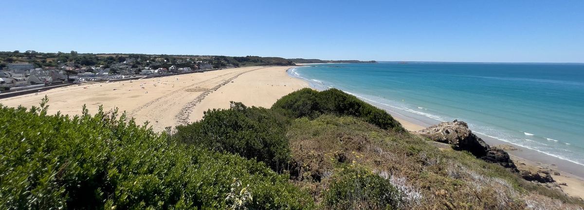 Erquy vue depuis la falaise : la longue plage de sable, le bourg en bord de route et l'eau turquoise, un jour d'été